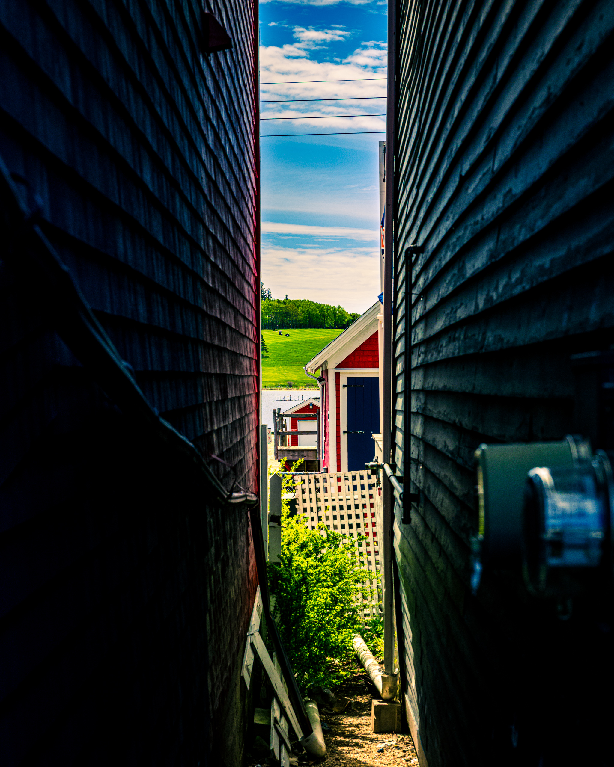 A narrow alleyway between two dark wooden buildings leads to a bright, open landscape. The alley is lined with a gravel path and some greenery. At the end, a red building with white trim stands against a backdrop of lush green fields, a waterway, and a clear blue sky with scattered clouds. Power lines stretch across the scene, adding to the rural setting.