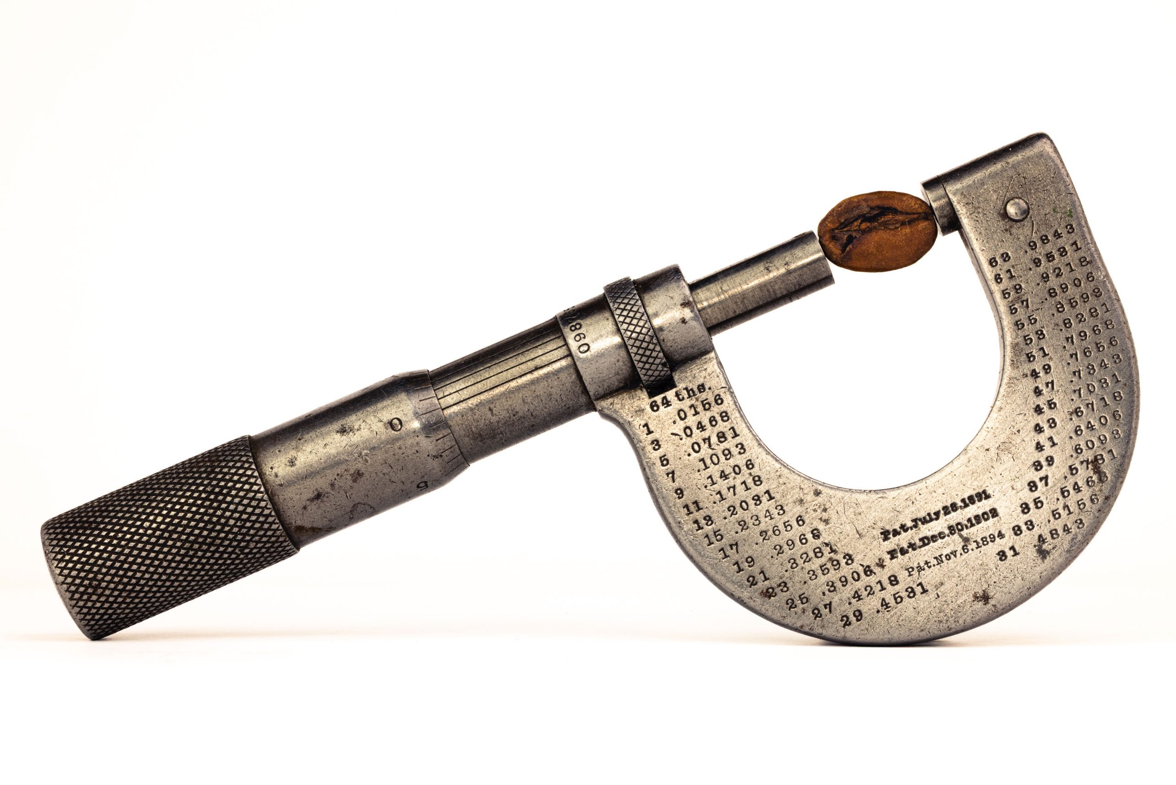 A vintage micrometer with a coffee bean held in its jaws, shown in profile against a white background. The C-frame body is engraved with a conversion table and three patent dates.