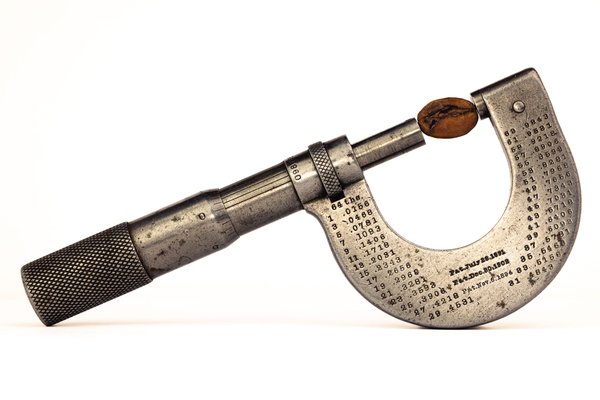 A vintage micrometer with a coffee bean held in its jaws, shown in profile against a white background. The C-frame body is engraved with a conversion table and three patent dates.