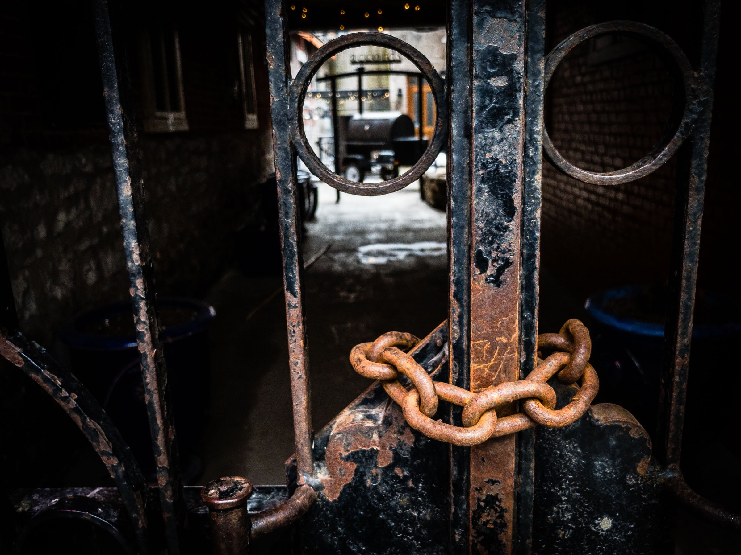 A rusted metal gate with circular designs blocks a dimly lit passageway. A thick, corroded chain wraps around the gate, securing it. Beyond the gate, a glimpse of a courtyard shows a brick wall, a blue container, and a partially visible grill or smoker on a trailer. The scene conveys a sense of secrecy and limited access to a gathering place in a hidden spot, with shadows enhancing the aged, weathered appearance of the metal and brick surfaces.