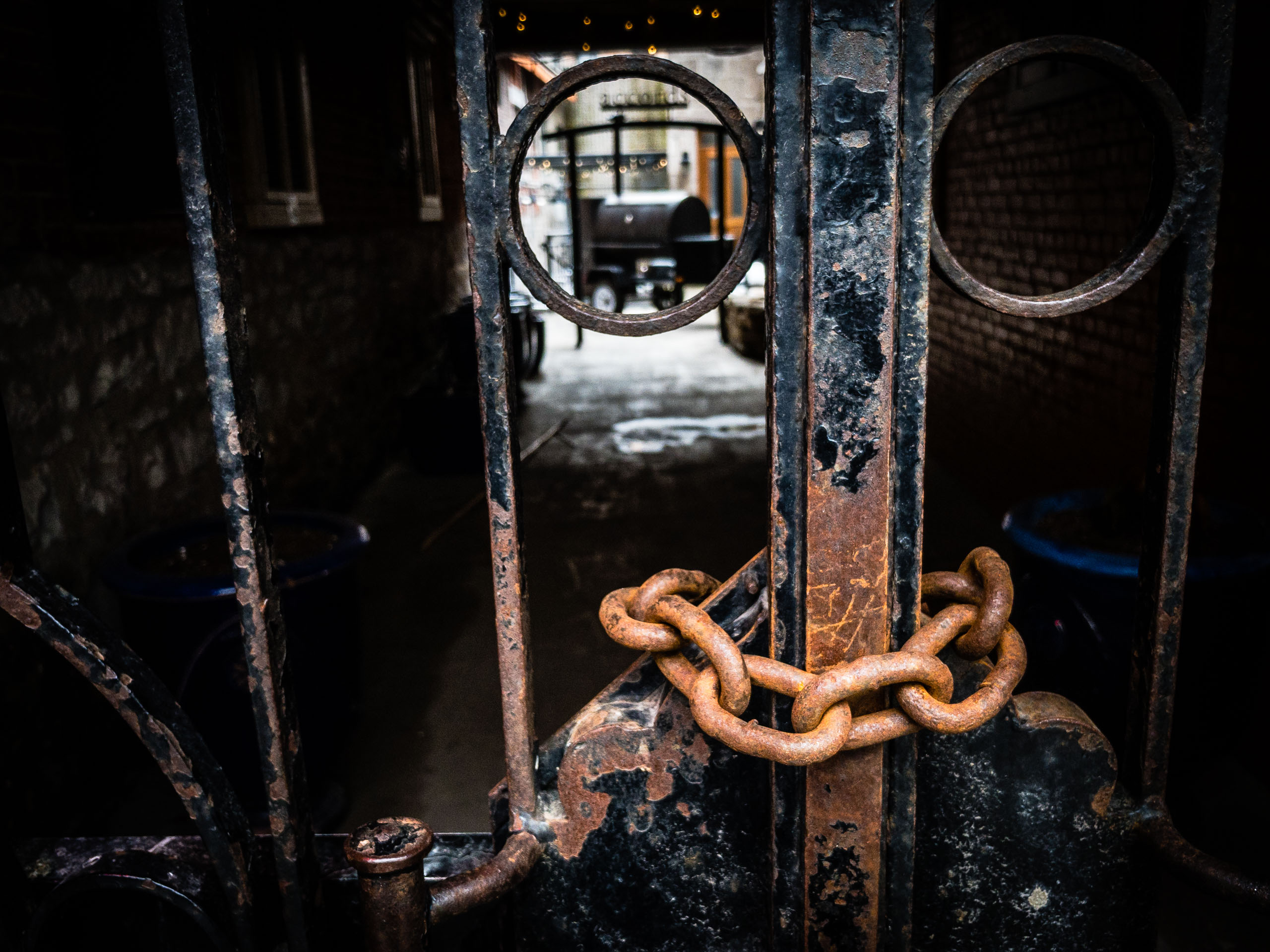 A rusted metal gate with circular designs blocks a dimly lit passageway. A thick, corroded chain wraps around the gate, securing it. Beyond the gate, a glimpse of a courtyard shows a brick wall, a blue container, and a partially visible grill or smoker on a trailer. The scene conveys a sense of secrecy and limited access to a gathering place in a hidden spot, with shadows enhancing the aged, weathered appearance of the metal and brick surfaces.