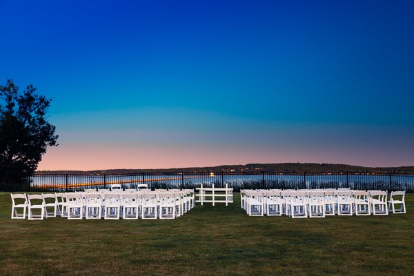 A serene outdoor wedding setup by the ocean at dusk. Rows of white folding chairs face a central aisle leading to a white arch. The sky transitions from deep blue to soft pink, casting a gentle glow over the scene. A black metal fence separates the grassy area from the calm water, with distant hills silhouetted against the horizon. Sparse trees frame the left side, adding to the tranquil atmosphere.