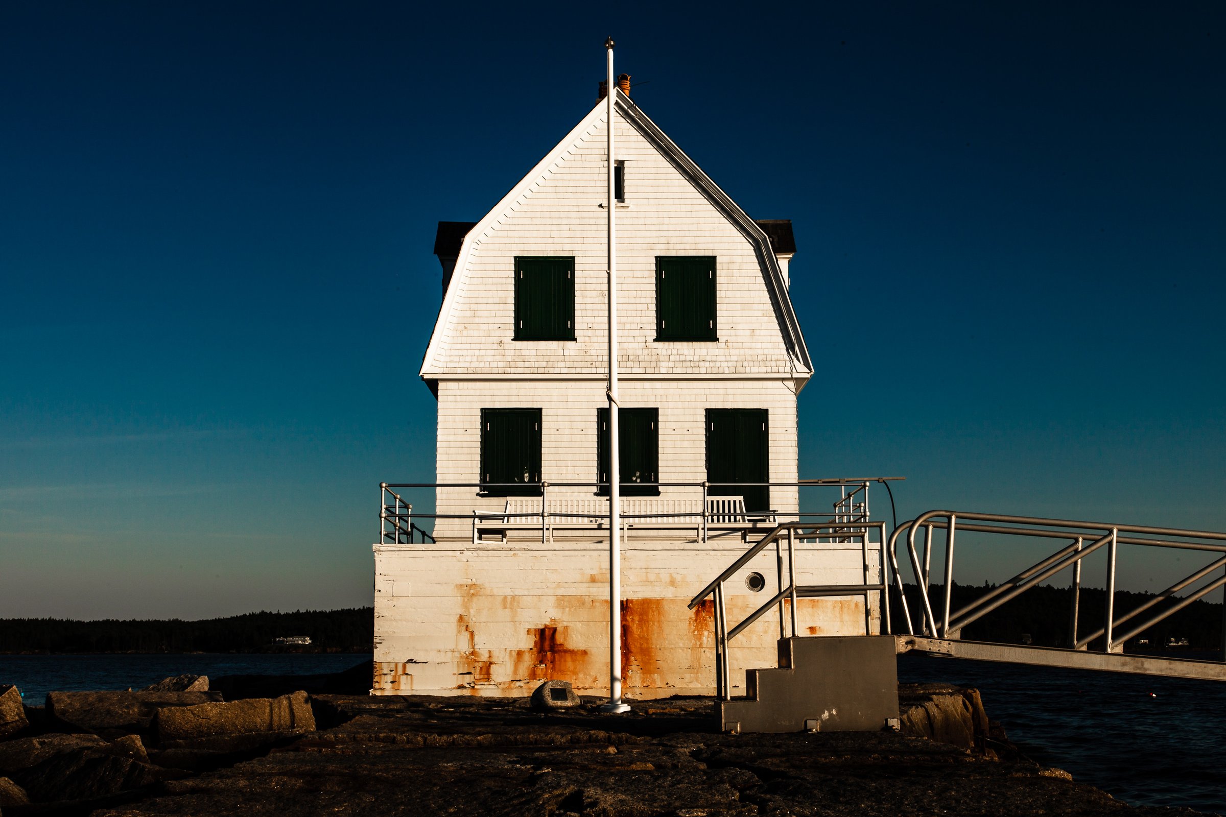 A white, three-story building with a steep roof stands by the water. The structure has dark green shutters and a weathered, rusty base. A metal ramp and railing lead up to it. The sky is a deep blue, transitioning to lighter shades near the horizon. Rocks and a small body of water surround the building, suggesting a coastal setting.