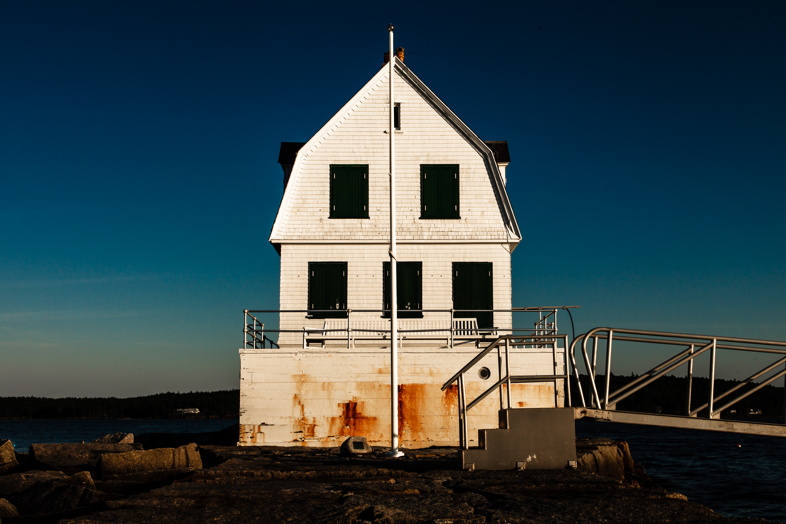 A white, three-story building with a steep roof stands by the water. The structure has dark green shutters and a weathered, rusty base. A metal ramp and railing lead up to it. The sky is a deep blue, transitioning to lighter shades near the horizon. Rocks and a small body of water surround the building, suggesting a coastal setting.