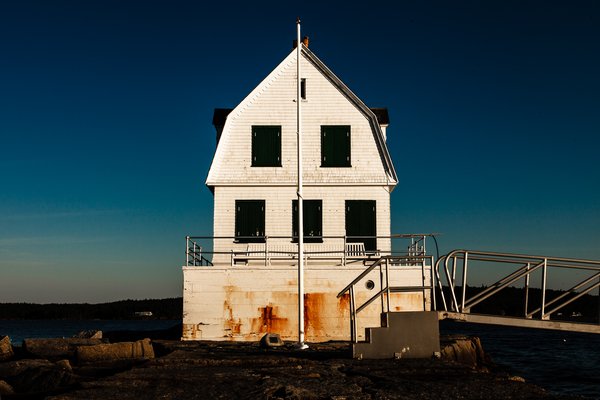 A white, three-story building with a steep roof stands by the water. The structure has dark green shutters and a weathered, rusty base. A metal ramp and railing lead up to it. The sky is a deep blue, transitioning to lighter shades near the horizon. Rocks and a small body of water surround the building, suggesting a coastal setting.