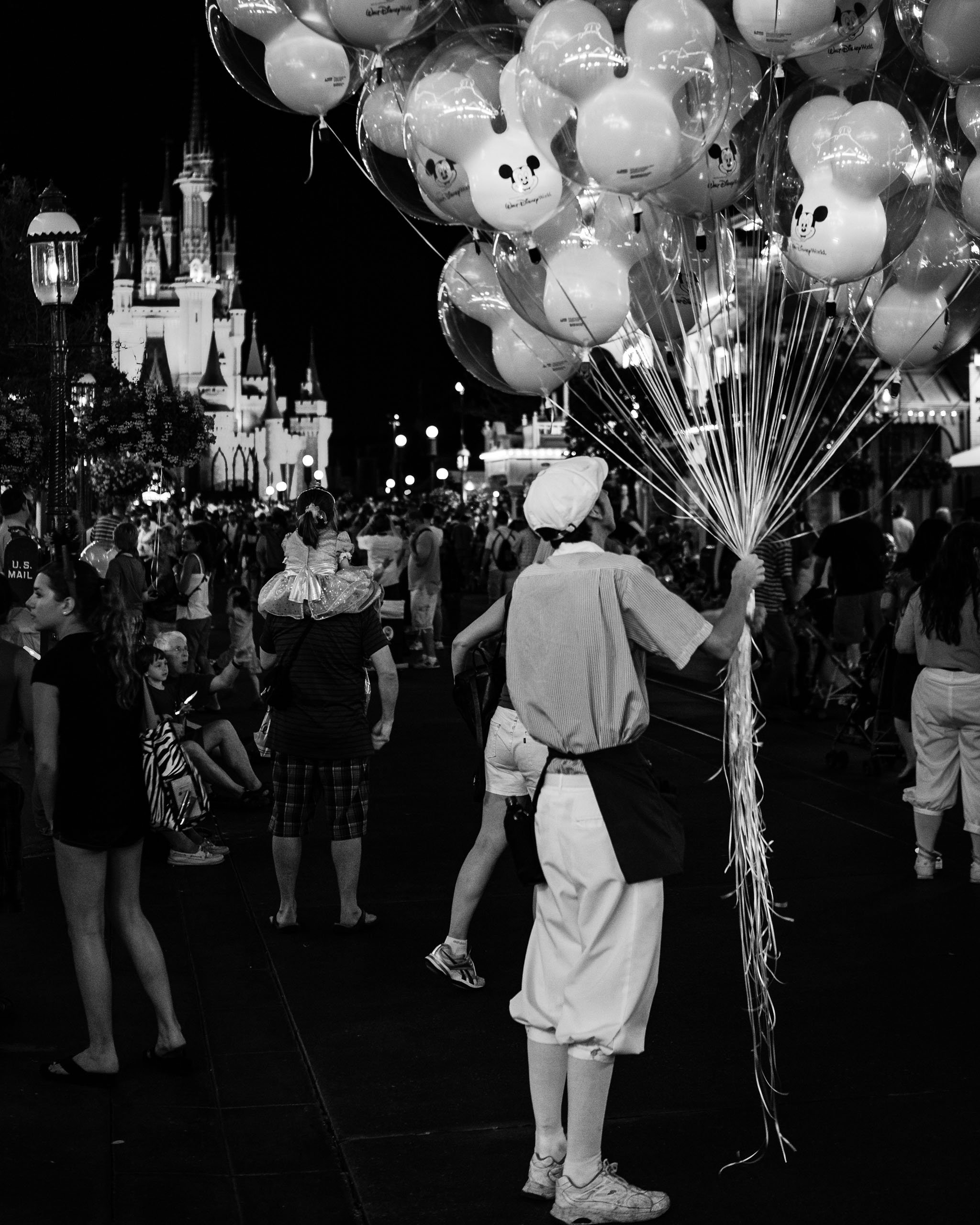 A person in a white hat and light clothing holds a large bunch of Mickey Mouse-shaped balloons in front of a brightly lit castle at night. The scene is bustling with people walking around, and the castle's spires are illuminated, creating a magical atmosphere. The balloons are clear with black Mickey faces. The overall mood is lively and enchanting, capturing the essence of a visit to Walt Disney World.