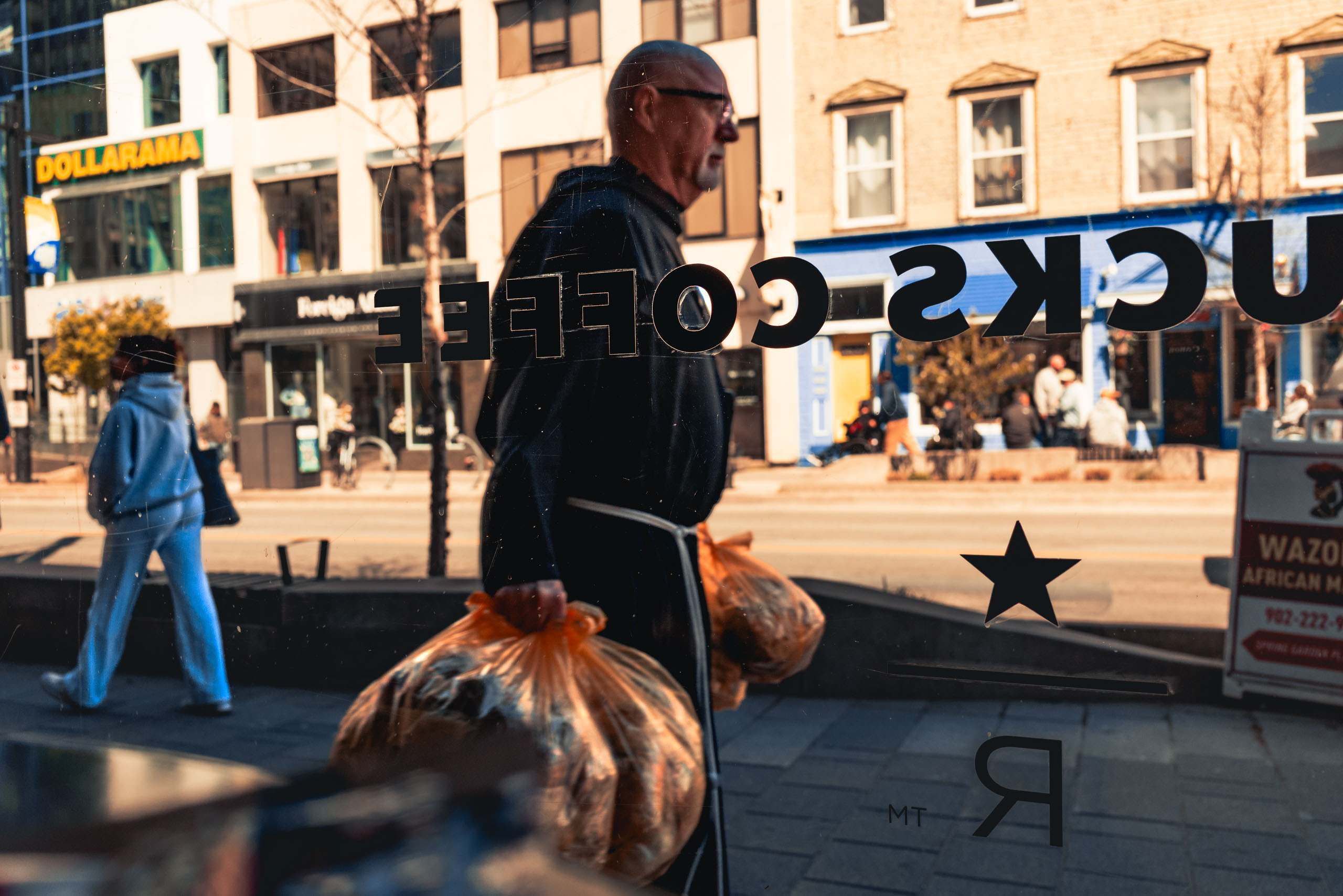 A man walks past a coffee shop window, holding two orange plastic bags filled with items. He has a grey goatee, wears a dark frock, likely religious in nature, and glasses. The background shows an empty street with pedestrians on adjacent walkways and other storefronts. The scene is lit by sunlight, casting shadows. Text and a star symbol is visible on the glass of the window through which the scene is viewed. The man walks in front of thr word 'COFFEE' in in the text on the window.