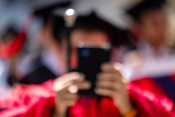 A person in a red graduation gown and cap holds a smartphone in front of their face, likely taking a photo. The background is blurred, showing other graduates in similar attire, suggesting a graduation ceremony. The lighting is bright, possibly from overhead lights, creating a festive atmosphere. The focus is on the smartphone and the person's hands, with the rest of the scene softly out of focus.