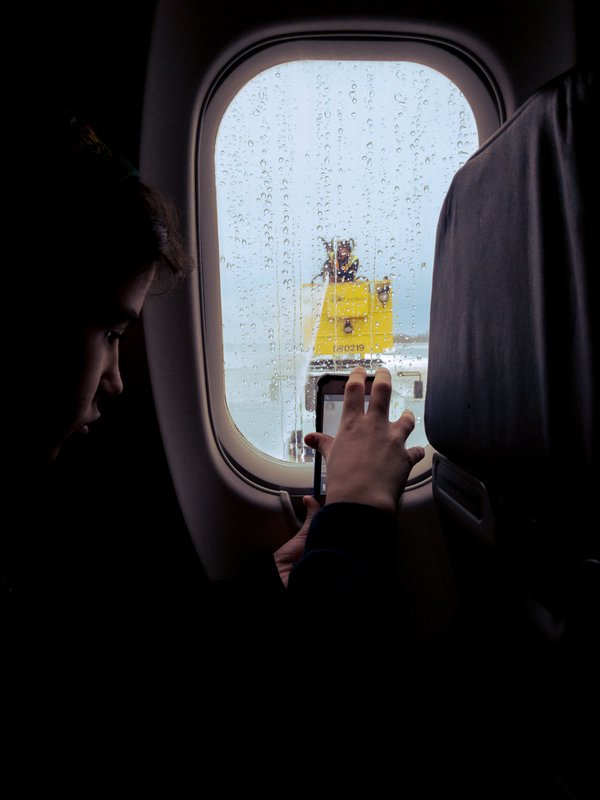 A person inside an airplane is taking a photo through a window covered in deicing fluid. Outside, a yellow deicing vehicle is visible on the tarmac, with a worker spraying the fluid on the plane and window. The worker is blurred by the presence of the fluid on the window. The scene is dimly lit, suggesting low light inside the airplane. The person's silhouette is partially visible, focused on capturing the moment with a smartphone.