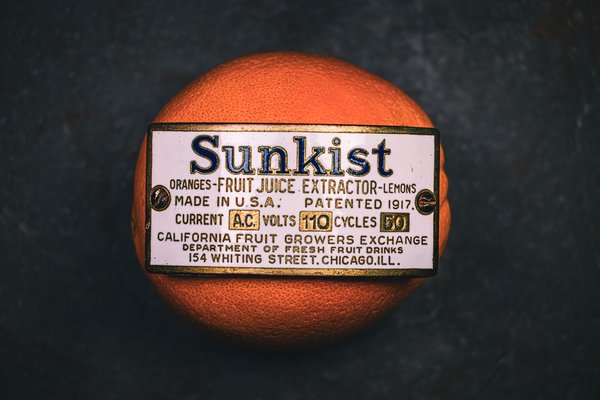 An orange with a metal patent marking plate from a vintage machine attached to it. The label reads "Sunskist" in bold blue letters, indicating the patent marking plate is for a Sunkist brand fruit juice extractor. The marking plate references the extractor being made in the U.S.A., patented in 1917, and that it operated on 110 volts and 50 cycles. The marking plate also references the California Fruit Growers Exchange and provides an address in Chicago, Illinois. The orange is set against a dark background, highlighting its vibrant color. Overall, the scene provides an interesting contrast with the patent marking plate from the extractor maching being directly attached to a fruit, the orange, on which the machine, not visible in the photograph, would have operated.