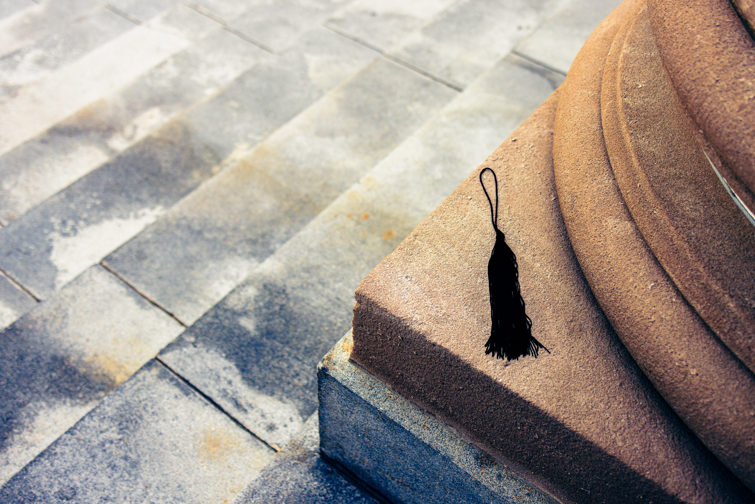 A black tassel lies on a stone pedestal of a column of a building. Large, rectangular gray stone steps with visible grout lines lead down and away from the column. The scene is well-lit, casting soft shadows on the stones. The tassel is the focal point, contrasting sharply with the muted tones of the stone and pavement.