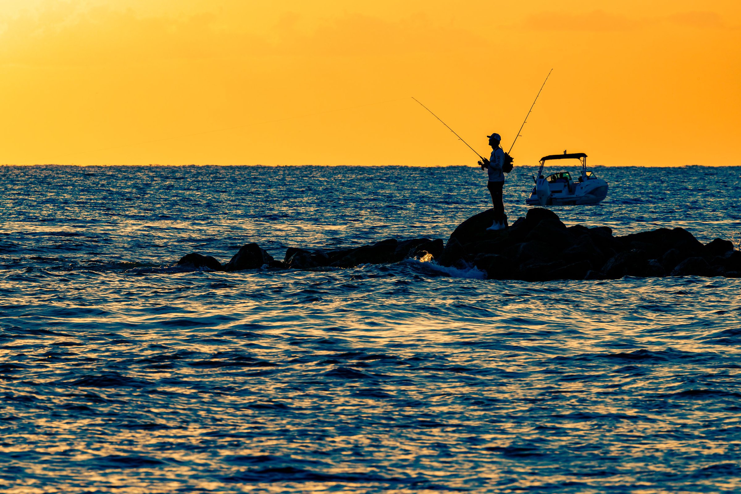 A lone angler stands in silhouette atop a rocky jetty extending into open water at sunset, two fishing rods arcing above him against a vivid sunset-drenched sky, with a motorboat anchored nearby in the deep blue water.