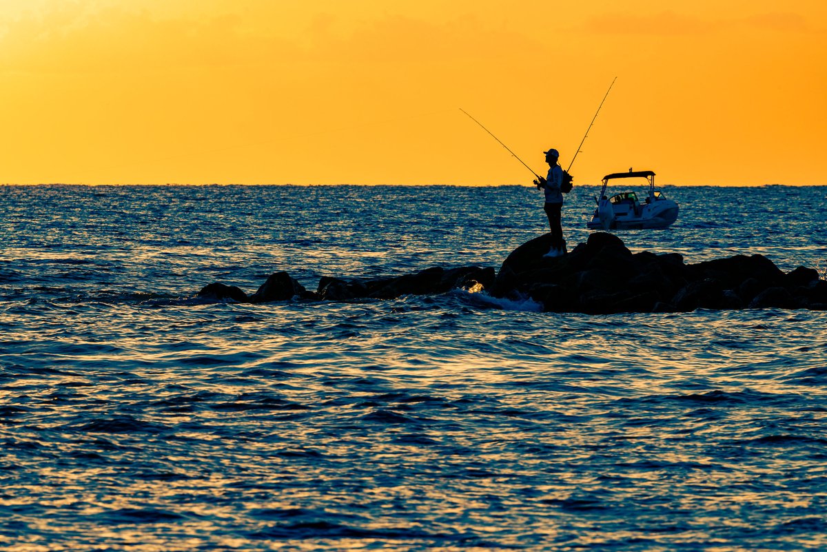 A lone angler stands in silhouette atop a rocky jetty extending into open water at sunset, two fishing rods arcing above him against a vivid sunset-drenched sky, with a motorboat anchored nearby in the deep blue water.