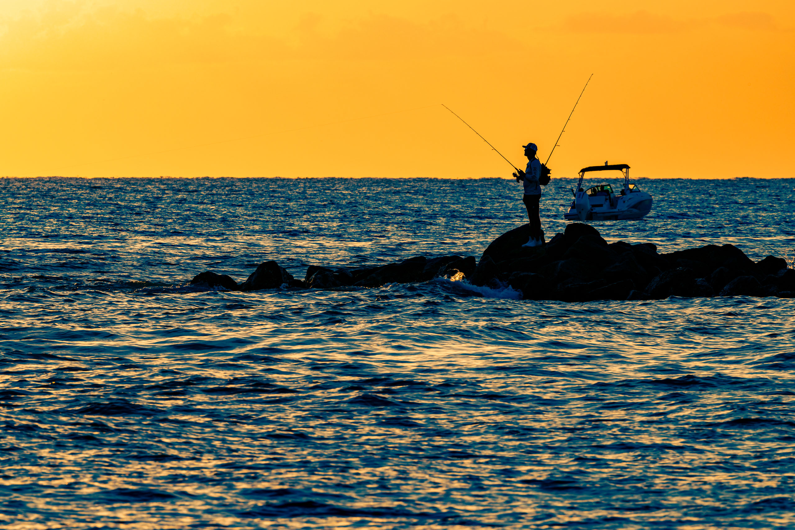 A lone angler stands in silhouette atop a rocky jetty extending into open water at sunset, two fishing rods arcing above him against a vivid sunset-drenched sky, with a motorboat anchored nearby in the deep blue water.