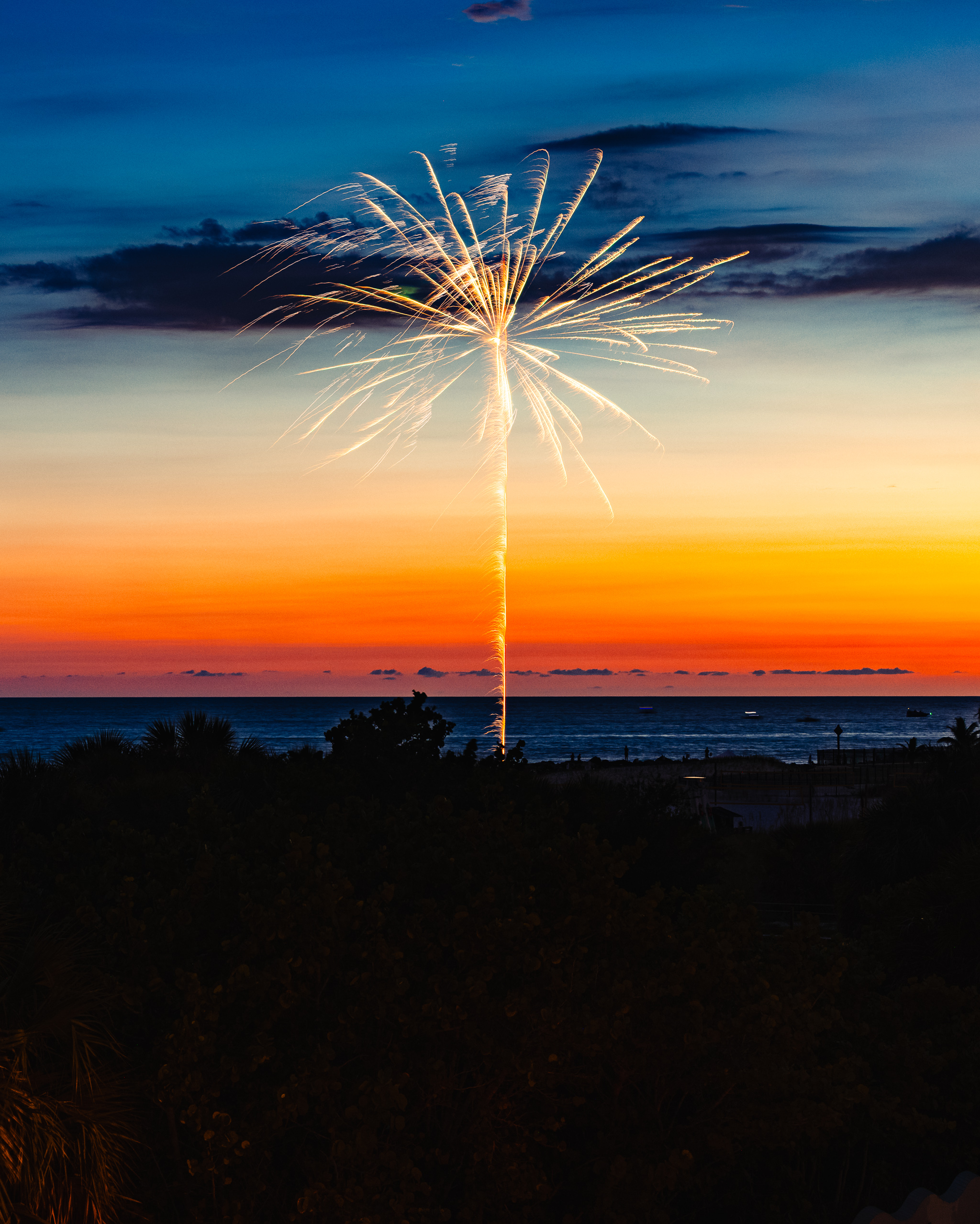 A vibrant sunset over the ocean, with deep blue transitioning to fiery orange and yellow near the horizon. Silhouetted against the sky is a single firework bursting in bright white and gold, creating a striking contrast. In the foreground, dark outlines of palm trees and shrubs add depth. The calm sea reflects the colorful sky, enhancing the serene yet festive atmosphere.