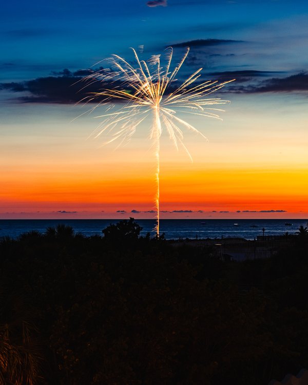 A vibrant sunset over the ocean, with deep blue transitioning to fiery orange and yellow near the horizon. Silhouetted against the sky is a single firework bursting in bright white and gold, creating a striking contrast. In the foreground, dark outlines of palm trees and shrubs add depth. The calm sea reflects the colorful sky, enhancing the serene yet festive atmosphere.