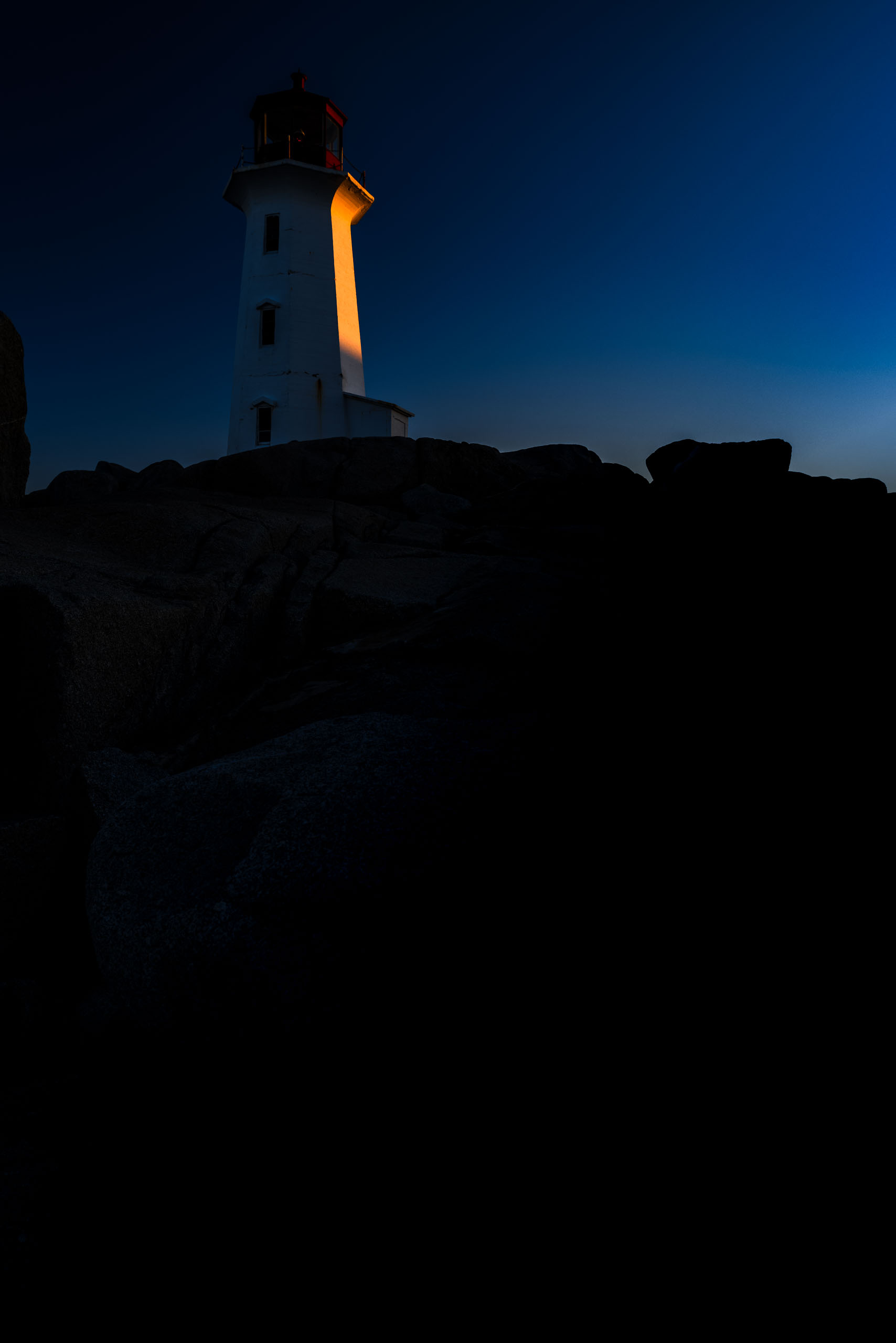 A white lighthouse stands atop rugged, dark rocks under a deep blue twilight sky. The light of sunrise glows warmly, casting a soft illumination on a side of the lighthouse and subtly revealing the red  top of the structure. The scene conveys a sense of calm and isolation, with the lighthouse serving as a solitary beacon against the vastness of the sky and sea.