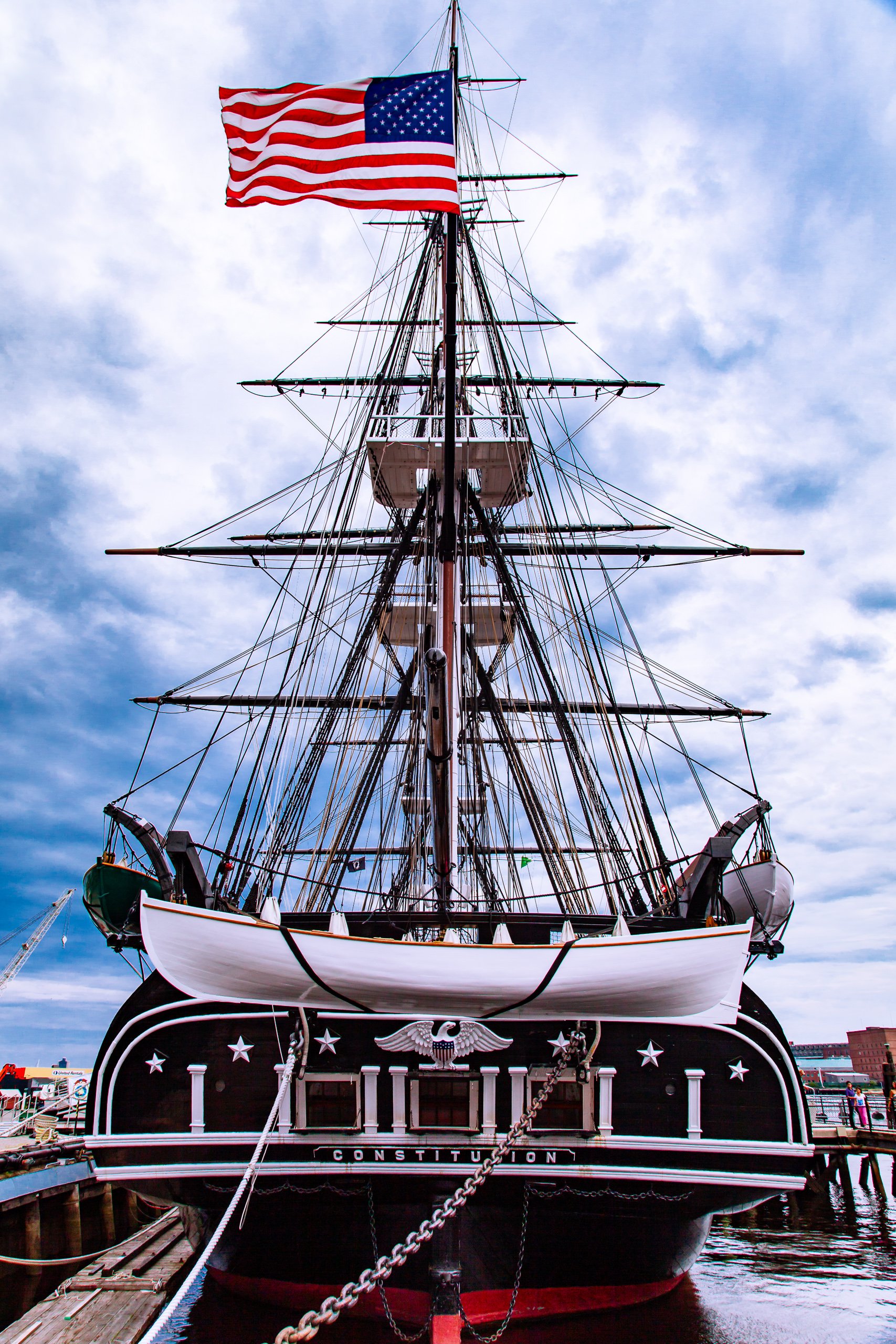 A tall, historic sailing ship with three masts flies an American flag at the top. The ship's hull is dark with white trim and features the name "Constitution" below an eagle emblem. The bow is ornate, and the ship is docked in calm water. The sky is partly cloudy, adding a dramatic backdrop to the scene.