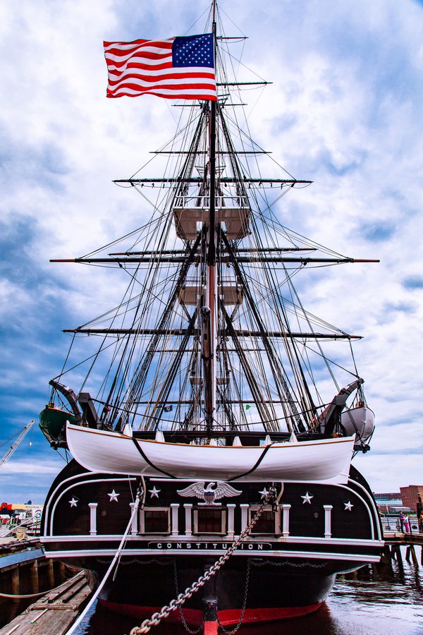 A tall, historic sailing ship with three masts flies an American flag at the top. The ship's hull is dark with white trim and features the name "Constitution" below an eagle emblem. The bow is ornate, and the ship is docked in calm water. The sky is partly cloudy, adding a dramatic backdrop to the scene.