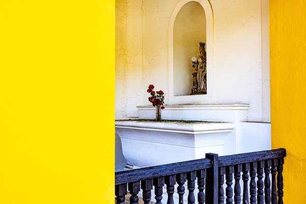 A small religious alcove in a wall features a statue of a robed figure holding a globe. Below the statue, a white pedestal holds a vase with red flowers. The alcove is set against a cream-colored wall with subtle cracks. To the left, a bright yellow wall contrasts sharply. A dark wooden railing with turned balusters runs along the bottom foreground, adding depth to the scene.