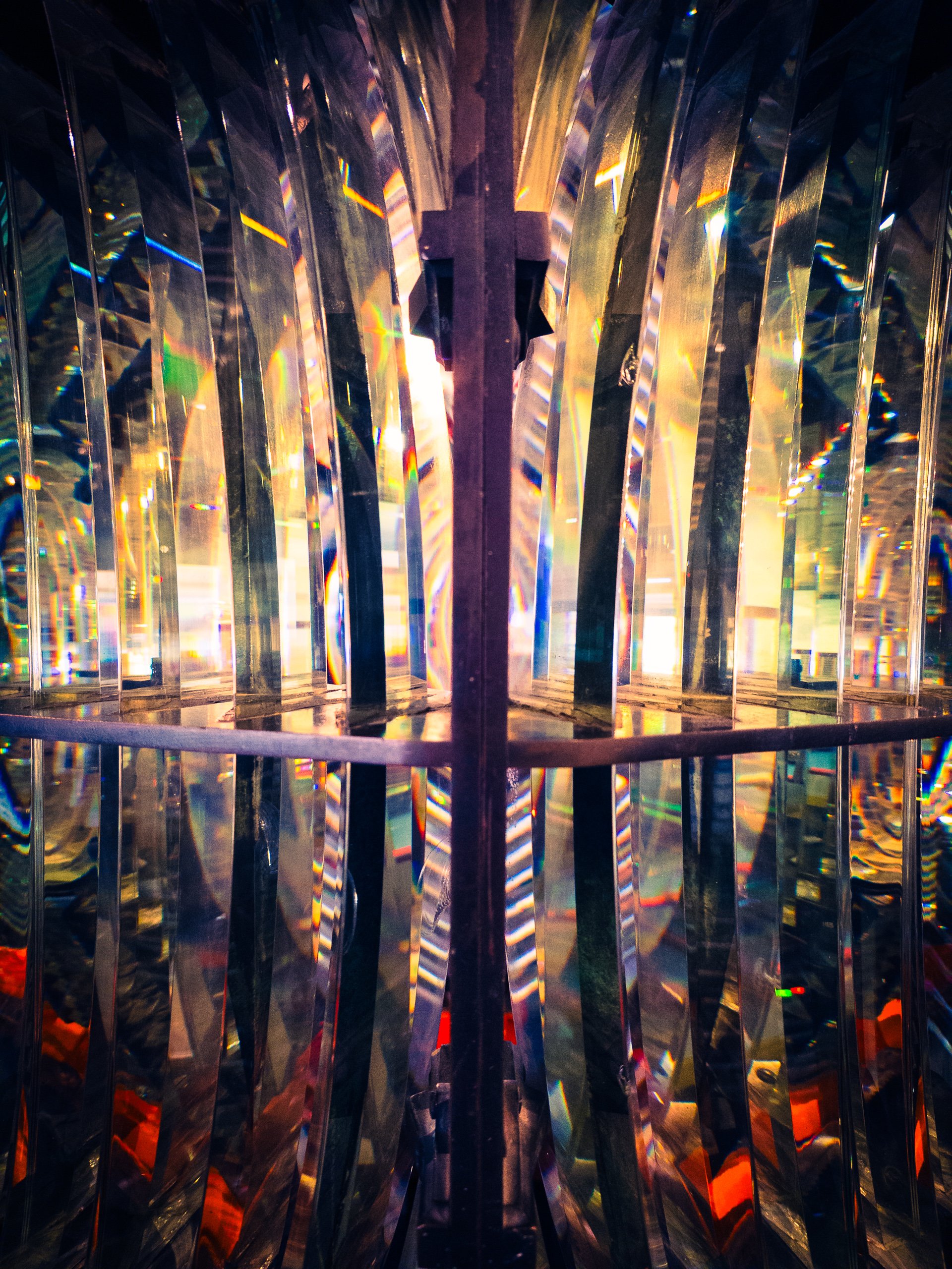 A kaleidoscope of colors and light filters through curved, transparent panels, creating a mesmerizing pattern. The panels are arranged in a radial symmetry, converging towards a central point. The light appears to be artificial, with hues of blue, red, yellow, and green blending seamlessly. The overall effect is abstract and dynamic, evoking a sense of movement and energy. The structure seems to be enclosed, possibly part of an art installation or architectural feature. The interplay of light and shadow adds depth and complexity to the scene, making it visually captivating and intriguing.
