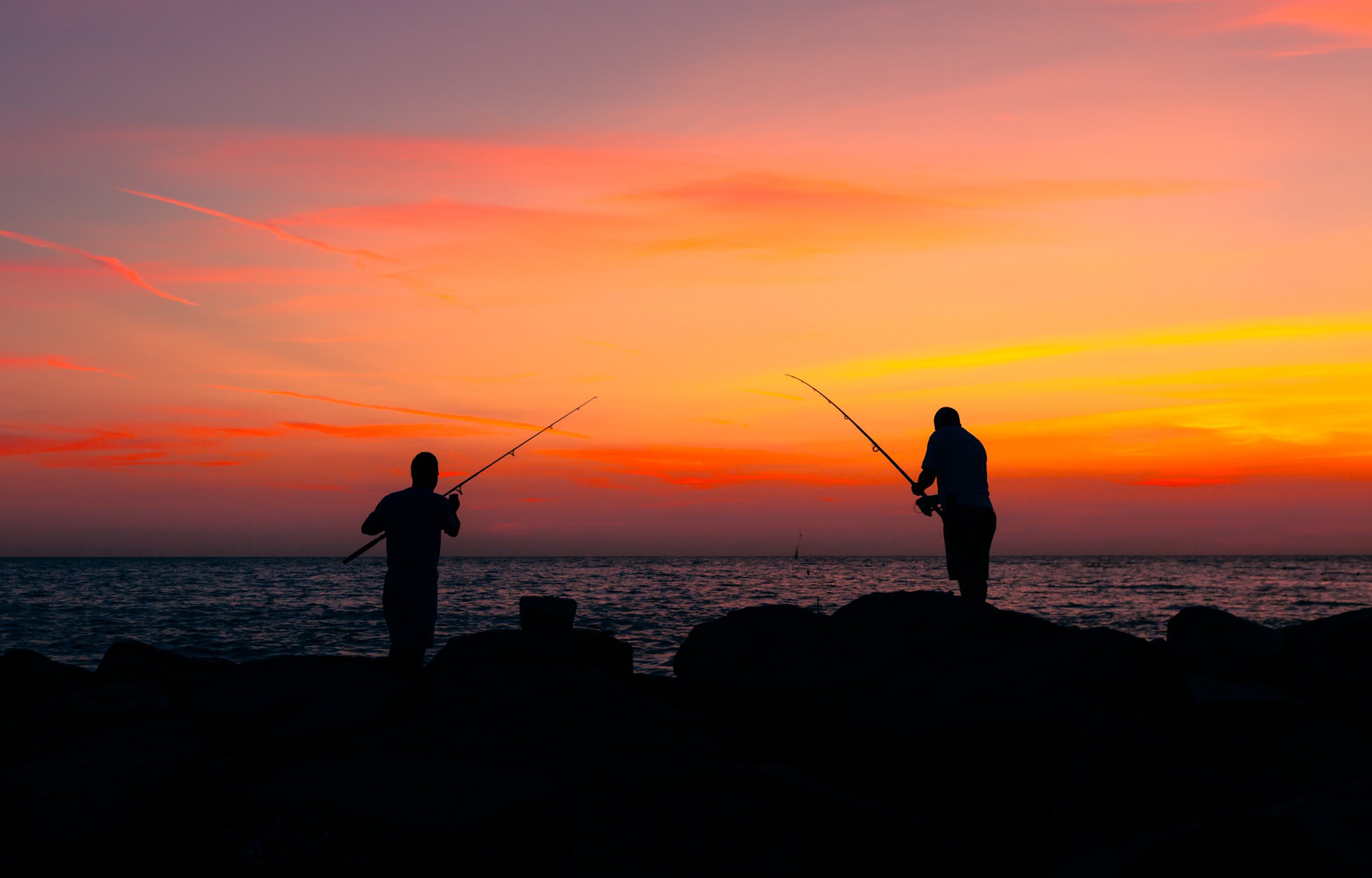 Two silhouetted men stand on a rocky pier by the ocean, fishing rods in hand. The sky is a vibrant gradient of orange, pink, and purple, indicating a sunset over the ocean. The calm ocean is dark, slightly reflecting the deepes colors of the sky, creating a serene atmosphere. The figures are positioned apart, each focused on their activity, with fishing rods pointed toward each other, embodying a peaceful moment of solitude and connection with nature.