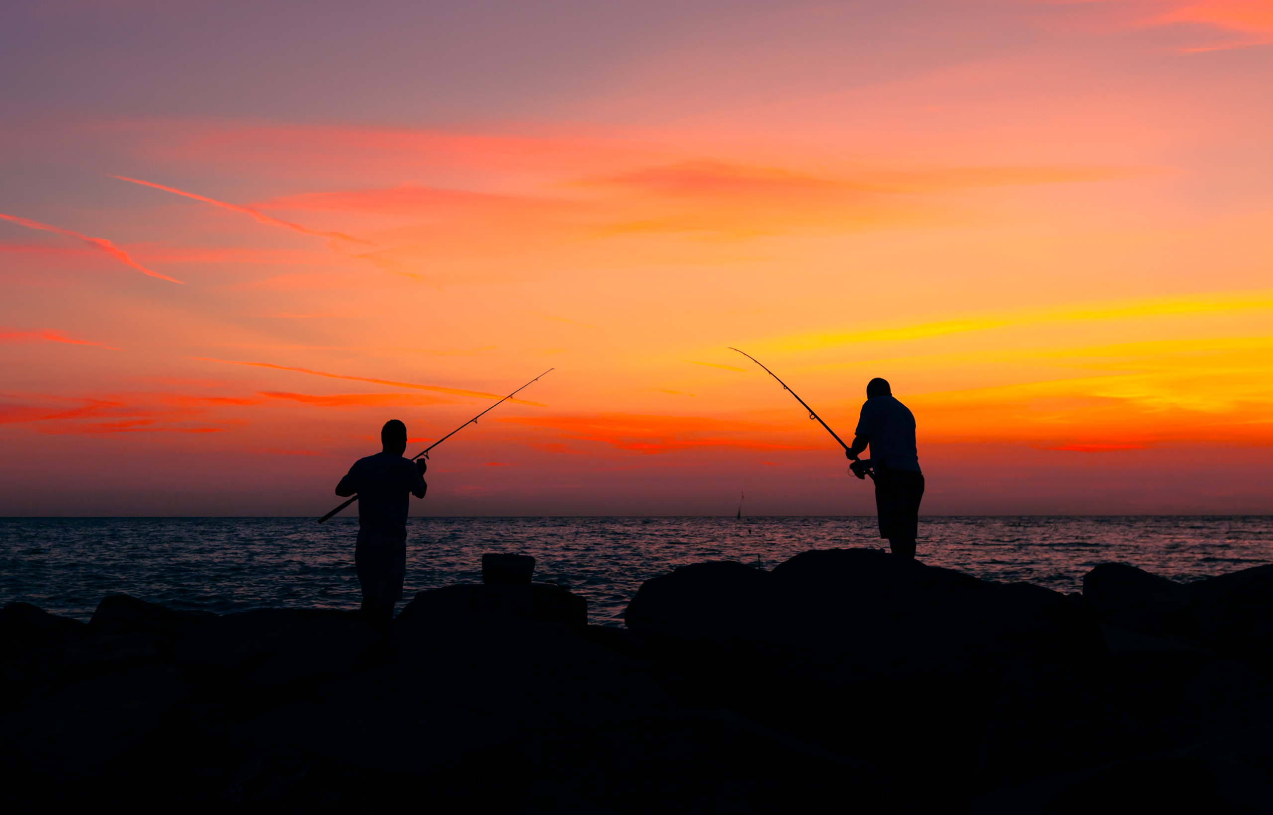 Two silhouetted men stand on a rocky pier by the ocean, fishing rods in hand. The sky is a vibrant gradient of orange, pink, and purple, indicating a sunset over the ocean. The calm ocean is dark, slightly reflecting the deepes colors of the sky, creating a serene atmosphere. The figures are positioned apart, each focused on their activity, with fishing rods pointed toward each other, embodying a peaceful moment of solitude and connection with nature.