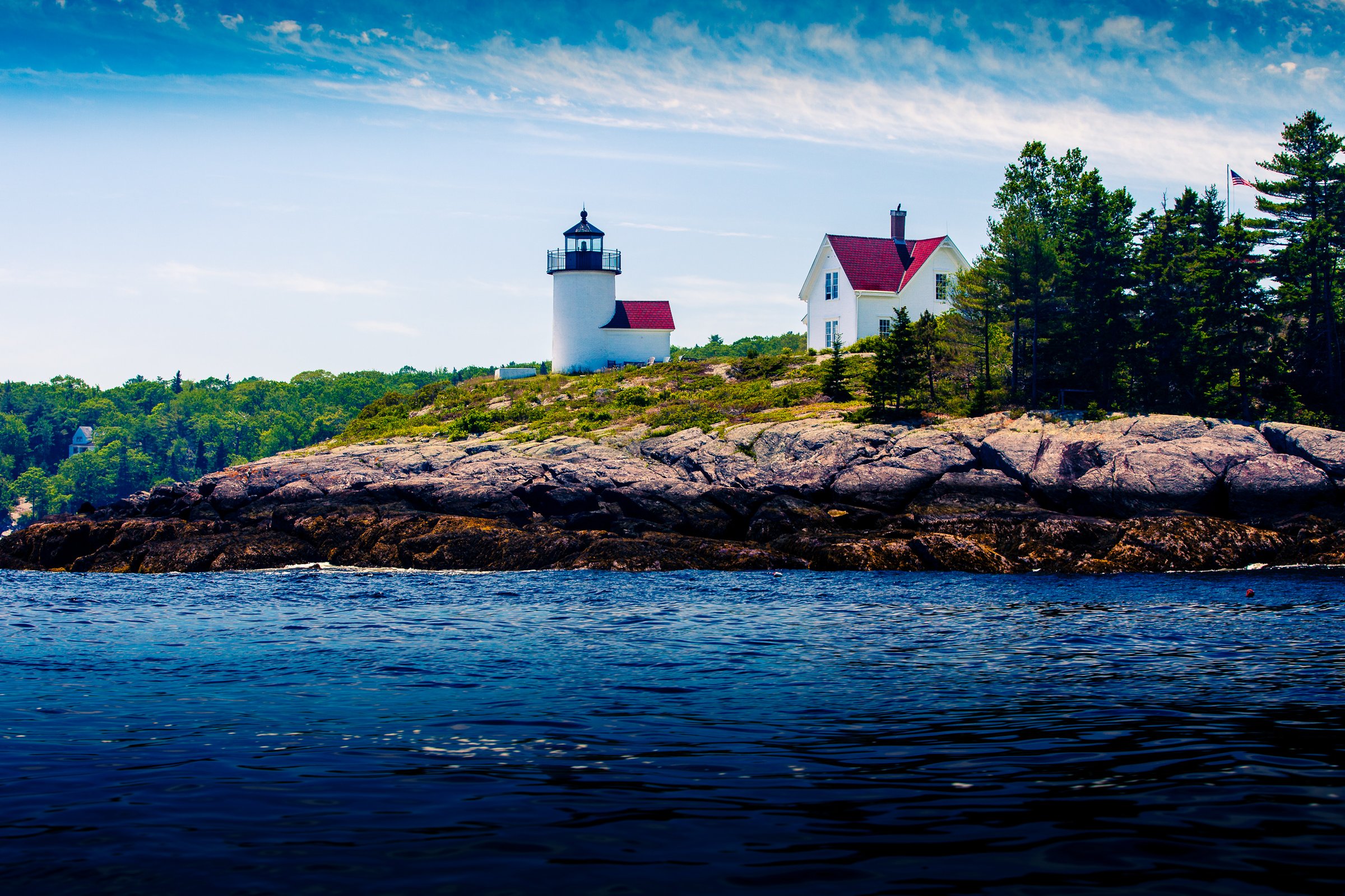 A picturesque coastal scene features a white lighthouse with a black top and red roof on a rocky outcrop. Adjacent is a white house with a red roof, surrounded by lush green trees. The rocky shoreline extends into a calm, deep blue sea under a bright sky with scattered white clouds. An American flag waves gently in the breeze, adding a touch of color and national pride to the serene landscape.