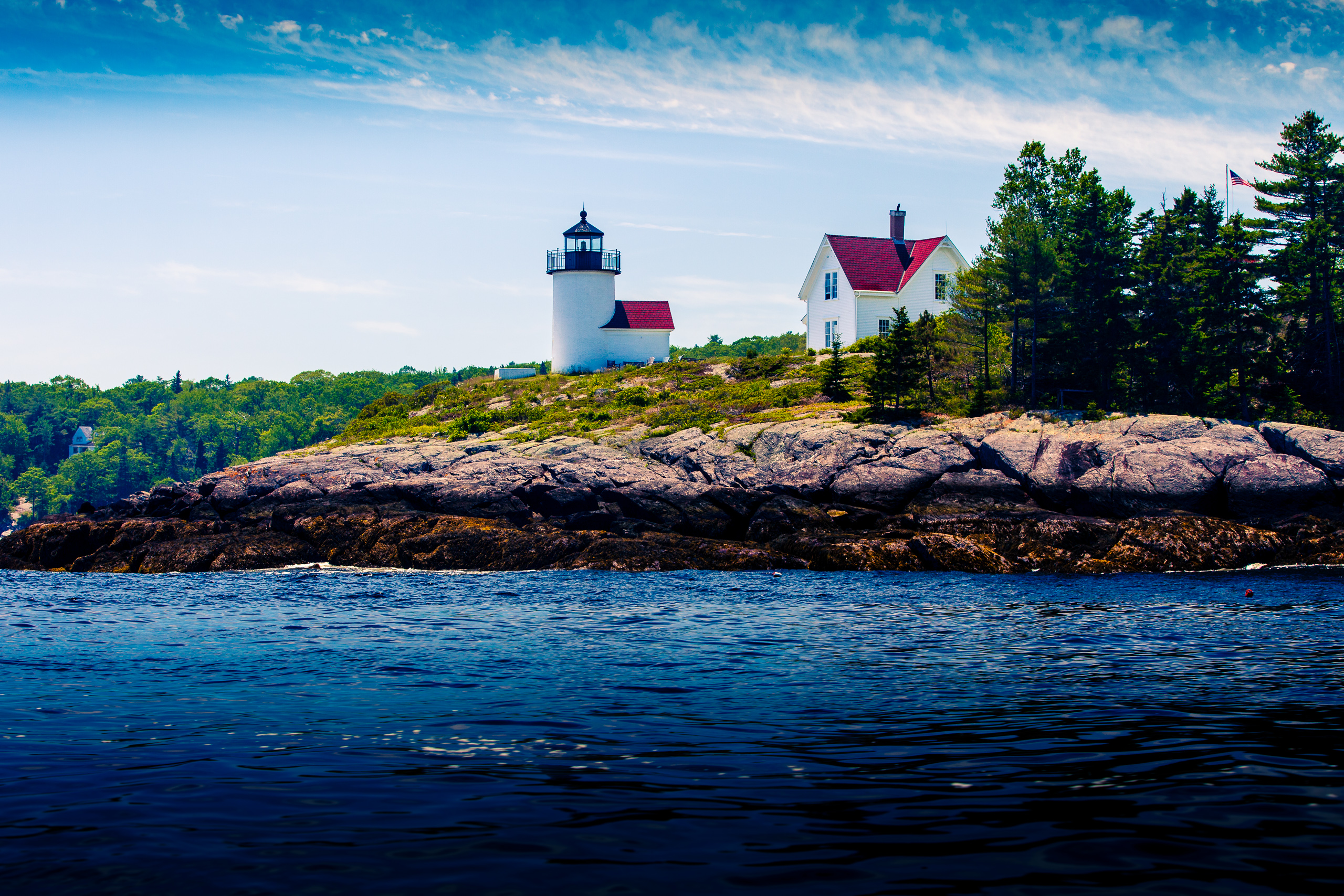 A picturesque coastal scene features a white lighthouse with a black top and red roof on a rocky outcrop. Adjacent is a white house with a red roof, surrounded by lush green trees. The rocky shoreline extends into a calm, deep blue sea under a bright sky with scattered white clouds. An American flag waves gently in the breeze, adding a touch of color and national pride to the serene landscape.