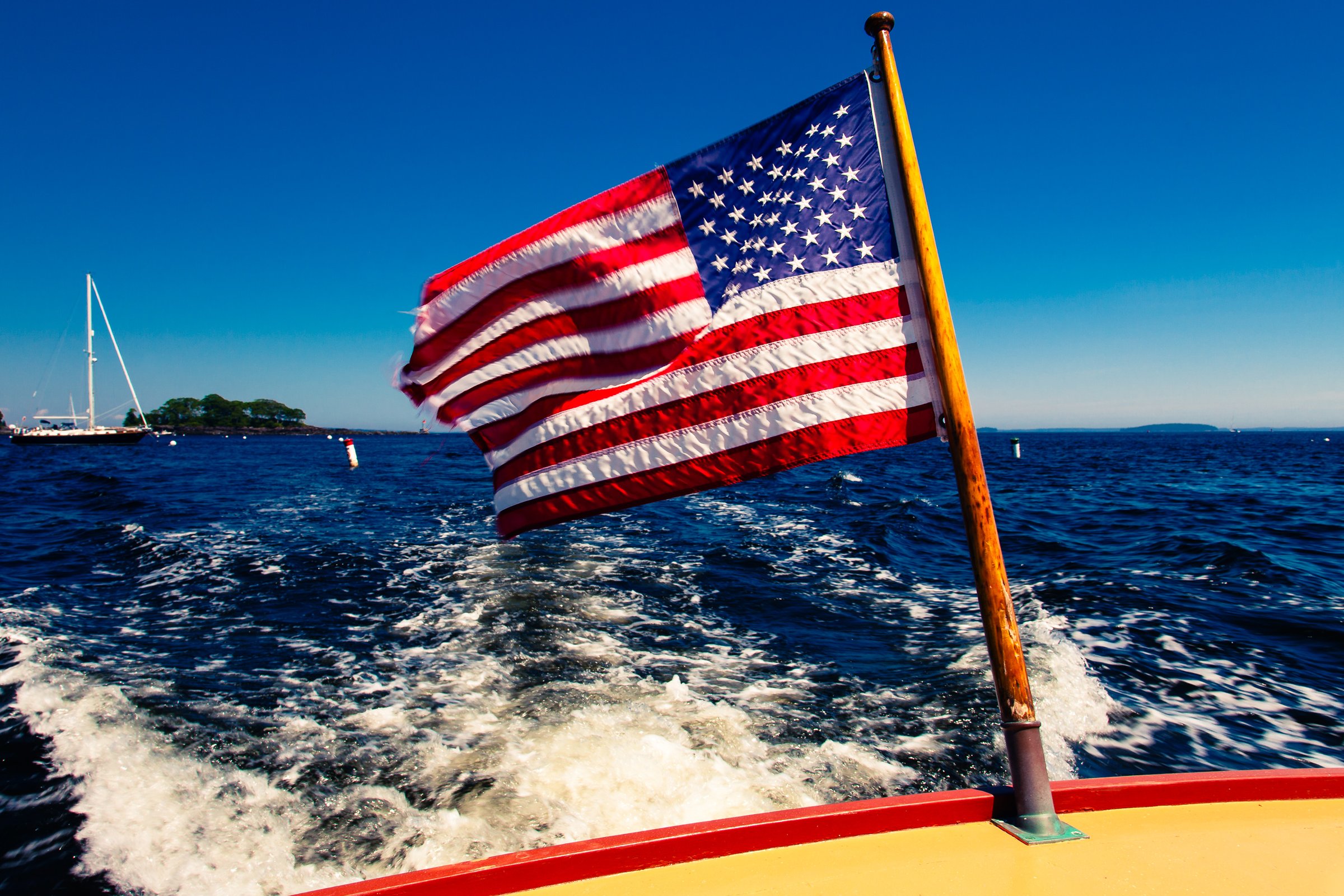 An American flag waves on a boat moving through deep blue water. The flag's red, white, and blue colors stand out against the clear sky. The boat's wake creates white foam beneath the flagpole. In the distance, a sailboat and a small, green island are visible under a bright blue sky.