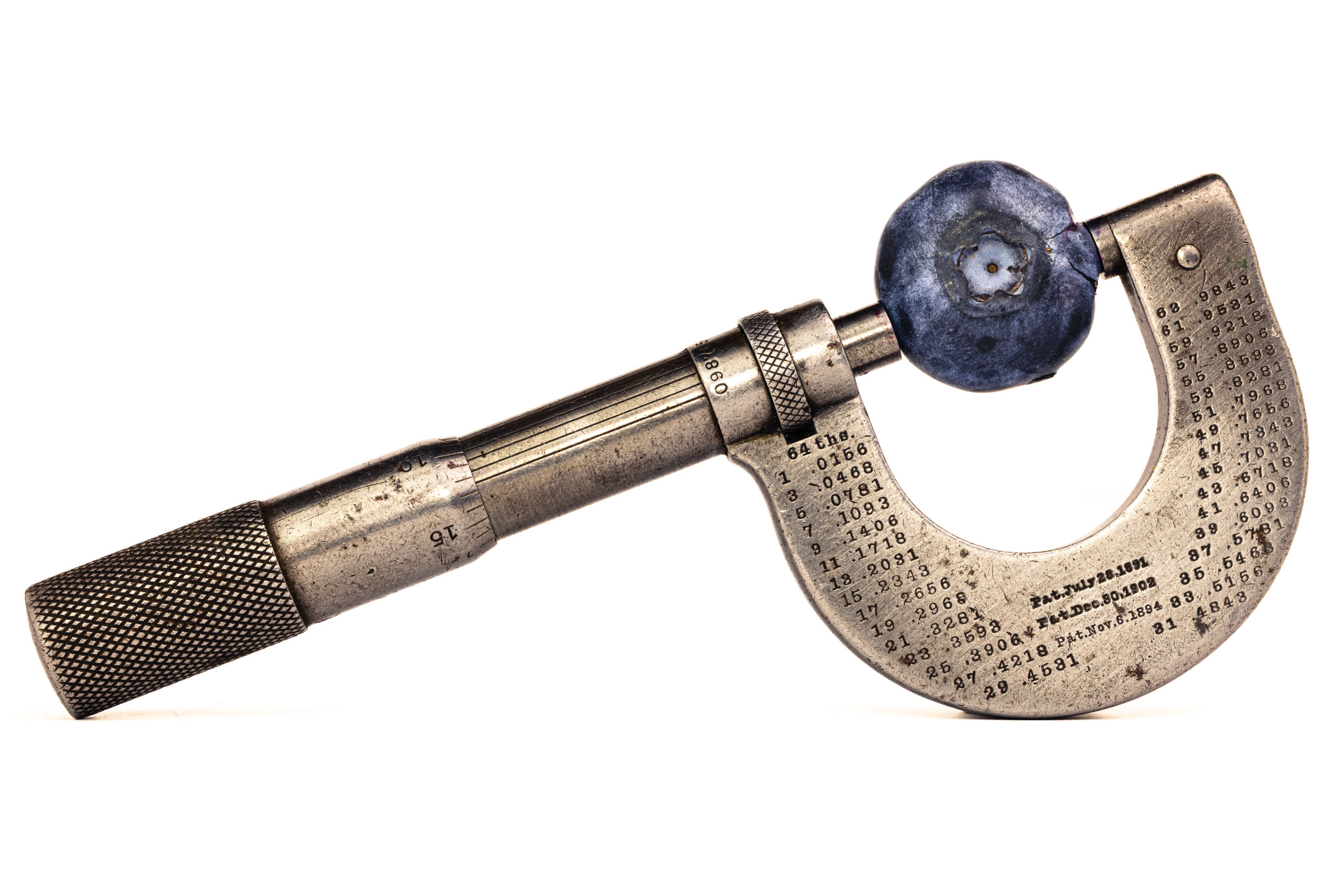 A vintage micrometer with a blueberry held in its jaws, shown in profile against a white background. The C-frame body is engraved with a conversion table and three patent dates.