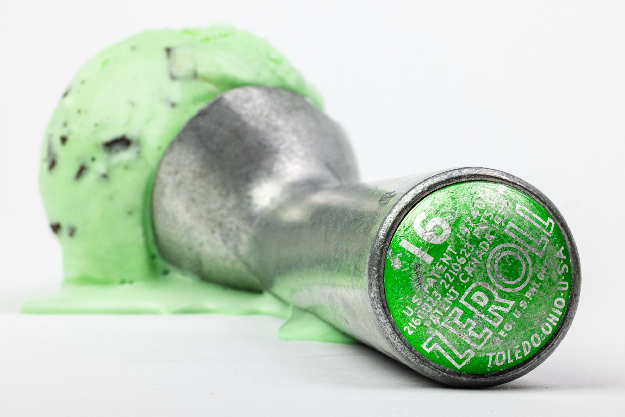 A vintage aluminum ice cream scoop with a patent-marked green disc reading "ZEROLL, Toledo, Ohio," resting in front of a melting scoop of mint chocolate chip ice cream.