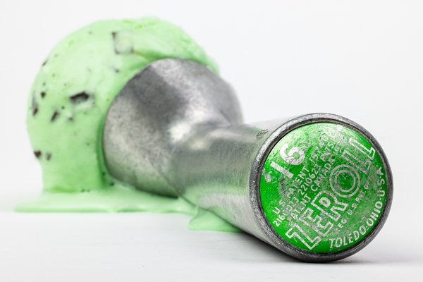 A vintage aluminum ice cream scoop with a patent-marked green disc reading "ZEROLL, Toledo, Ohio," resting in front of a melting scoop of mint chocolate chip ice cream.