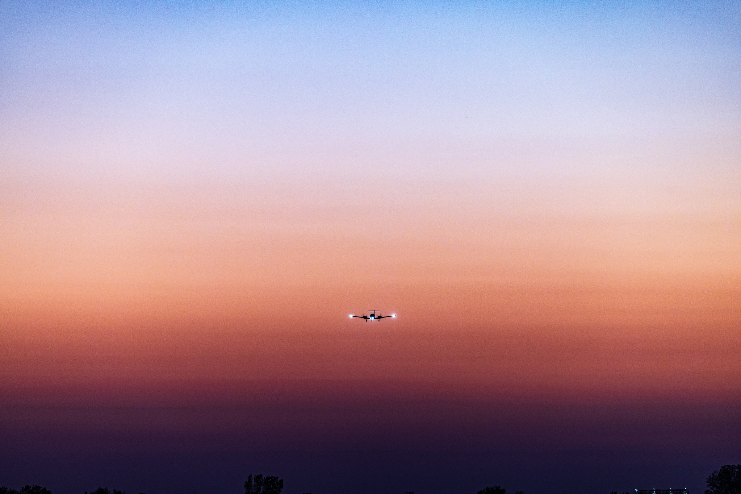 An airplane flies across a sky transitioning from deep blue at the top to warm orange and pink near the horizon. The silhouette of the aircraft is centered, with its lights visible against the colorful backdrop. Below, a dark line of trees forms the bottom edge of the scene, adding contrast to the vibrant sky.