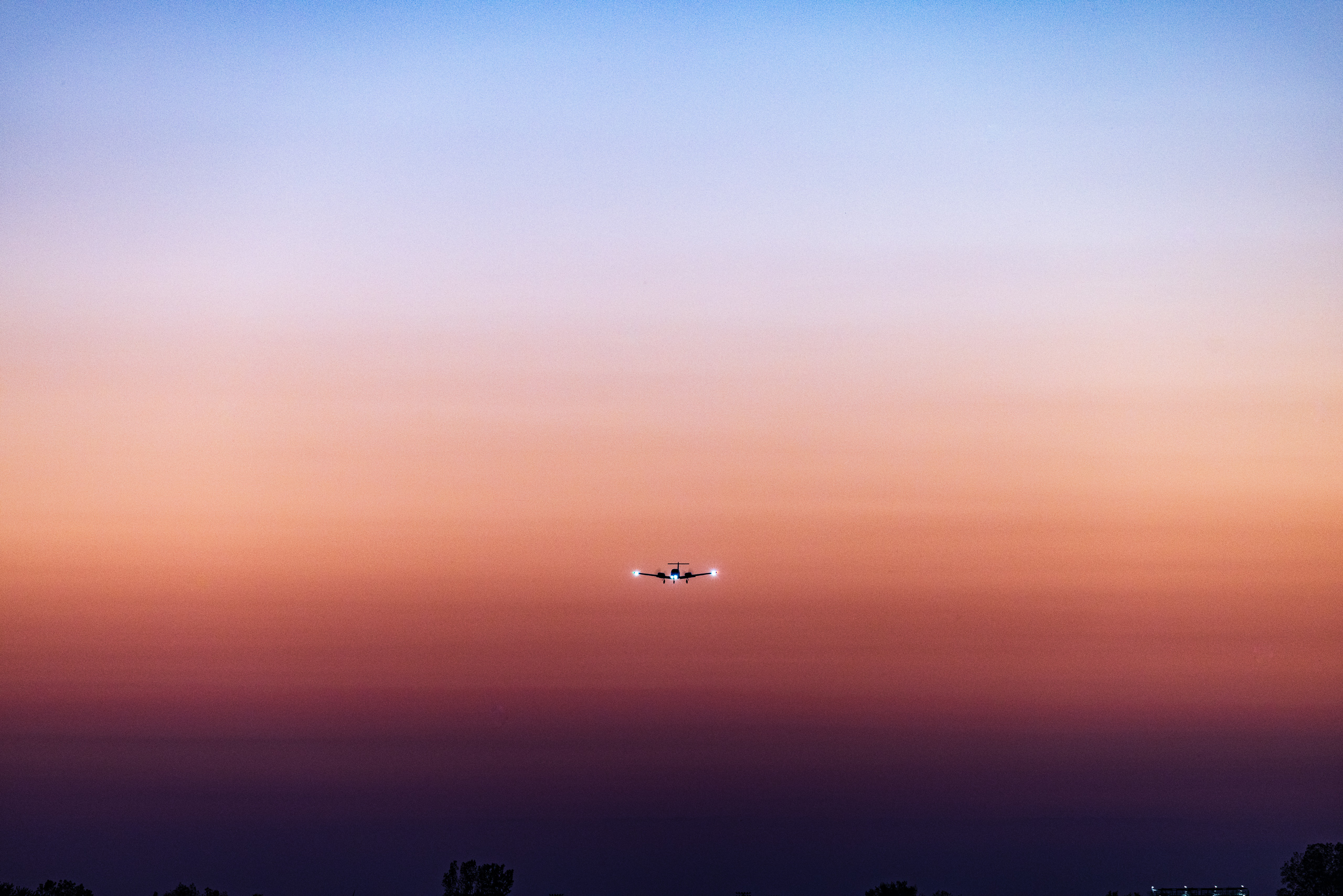 An airplane flies across a sky transitioning from deep blue at the top to warm orange and pink near the horizon. The silhouette of the aircraft is centered, with its lights visible against the colorful backdrop. Below, a dark line of trees forms the bottom edge of the scene, adding contrast to the vibrant sky.