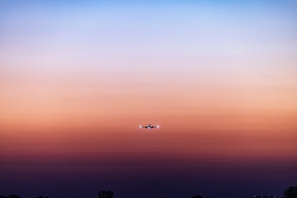 An airplane flies across a sky transitioning from deep blue at the top to warm orange and pink near the horizon. The silhouette of the aircraft is centered, with its lights visible against the colorful backdrop. Below, a dark line of trees forms the bottom edge of the scene, adding contrast to the vibrant sky.