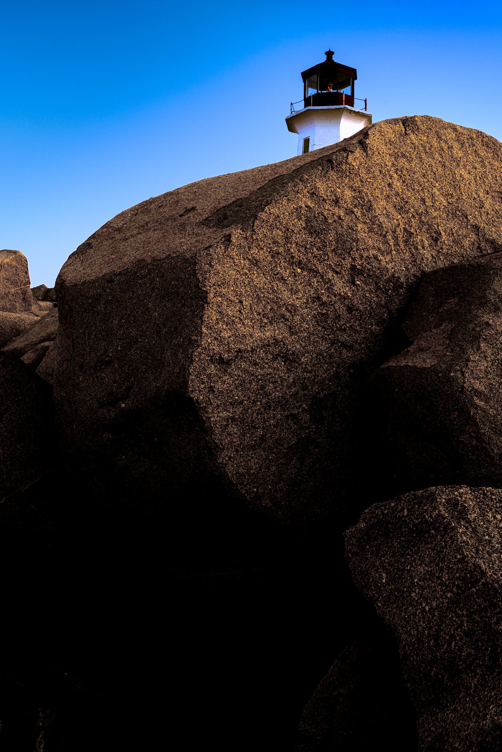 The top of a white lighthouse is visible over an edge of a large, smooth rock. The rock is bathed in warm sunlight, casting deep shadows. The sky above is a clear, vibrant blue, transitioning to a lighter hue near the horizon. The lighthouse stands prominently against the sky, its structure simple and functional. The scene conveys a sense of isolation and tranquility, typical of coastal landscapes.