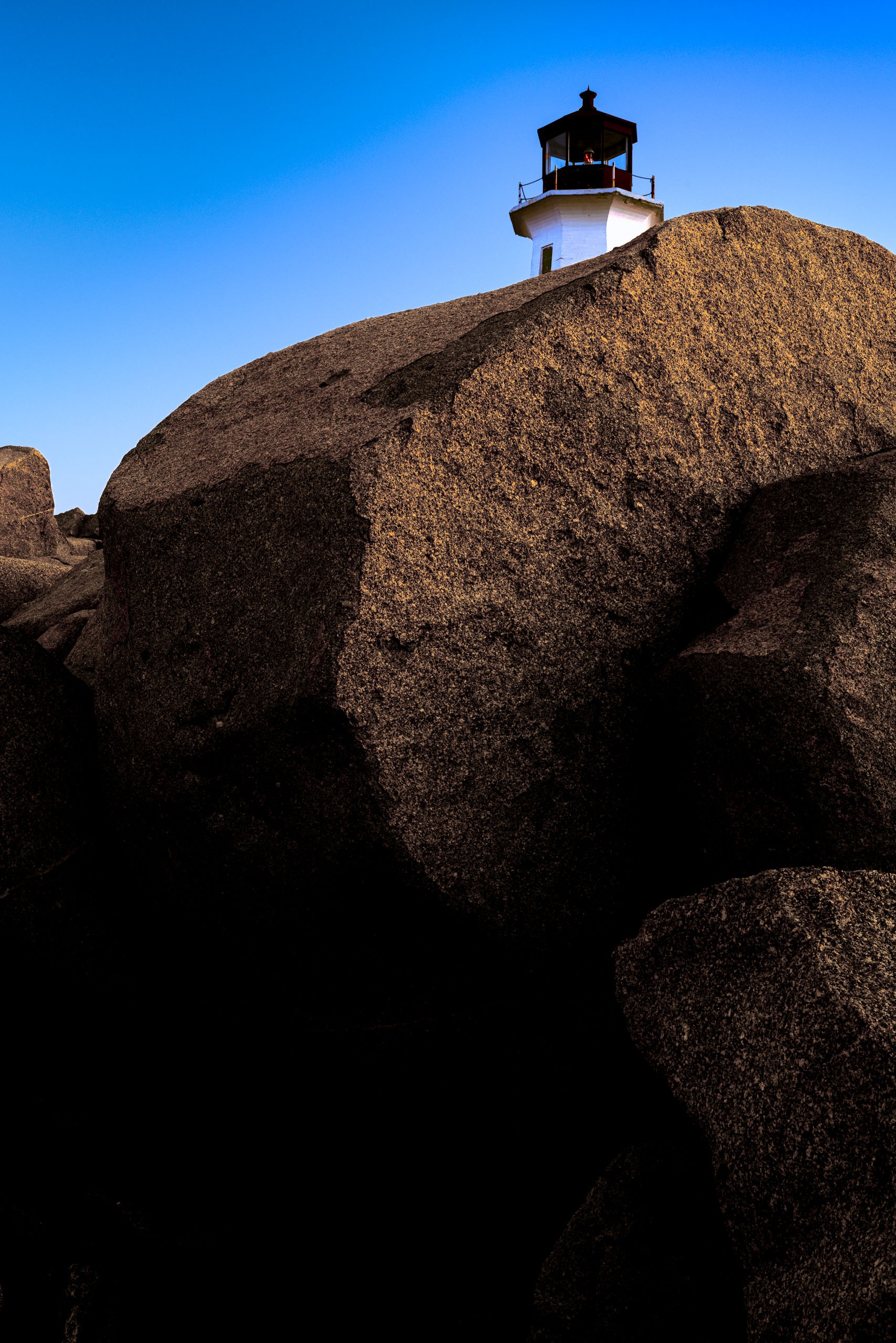The top of a white lighthouse is visible over an edge of a large, smooth rock. The rock is bathed in warm sunlight, casting deep shadows. The sky above is a clear, vibrant blue, transitioning to a lighter hue near the horizon. The lighthouse stands prominently against the sky, its structure simple and functional. The scene conveys a sense of isolation and tranquility, typical of coastal landscapes.