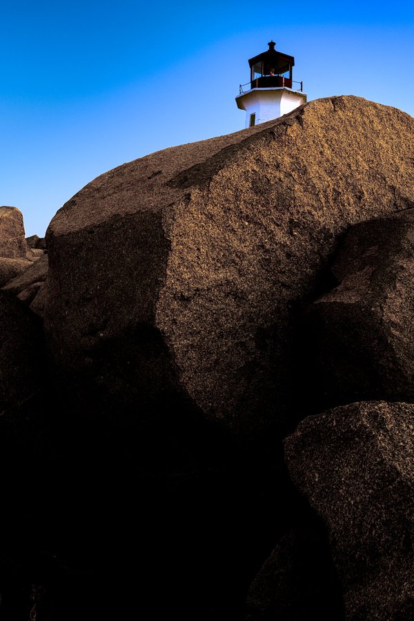 The top of a white lighthouse is visible over an edge of a large, smooth rock. The rock is bathed in warm sunlight, casting deep shadows. The sky above is a clear, vibrant blue, transitioning to a lighter hue near the horizon. The lighthouse stands prominently against the sky, its structure simple and functional. The scene conveys a sense of isolation and tranquility, typical of coastal landscapes.