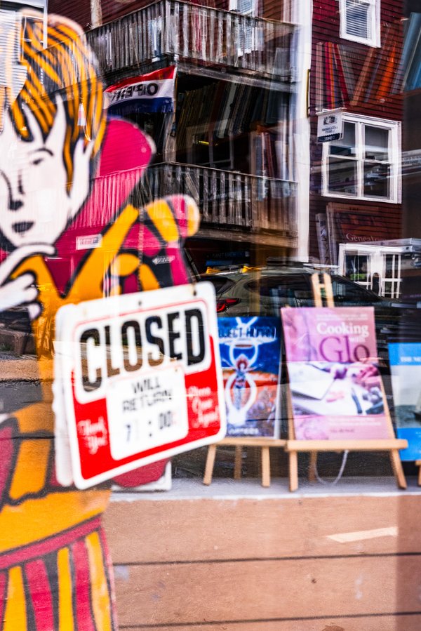 A storefront window displays a colorful cartoon character holding a microphone. A red and white "CLOSED" sign indicates the store is not open, with a note to return at 7:00. Behind the glass, various items are visible, including a blue box and a pink book titled "Cooking with Glo." The reflection of a brick building and parked cars can be seen on the window. The overall scene suggests a vibrant urban setting.