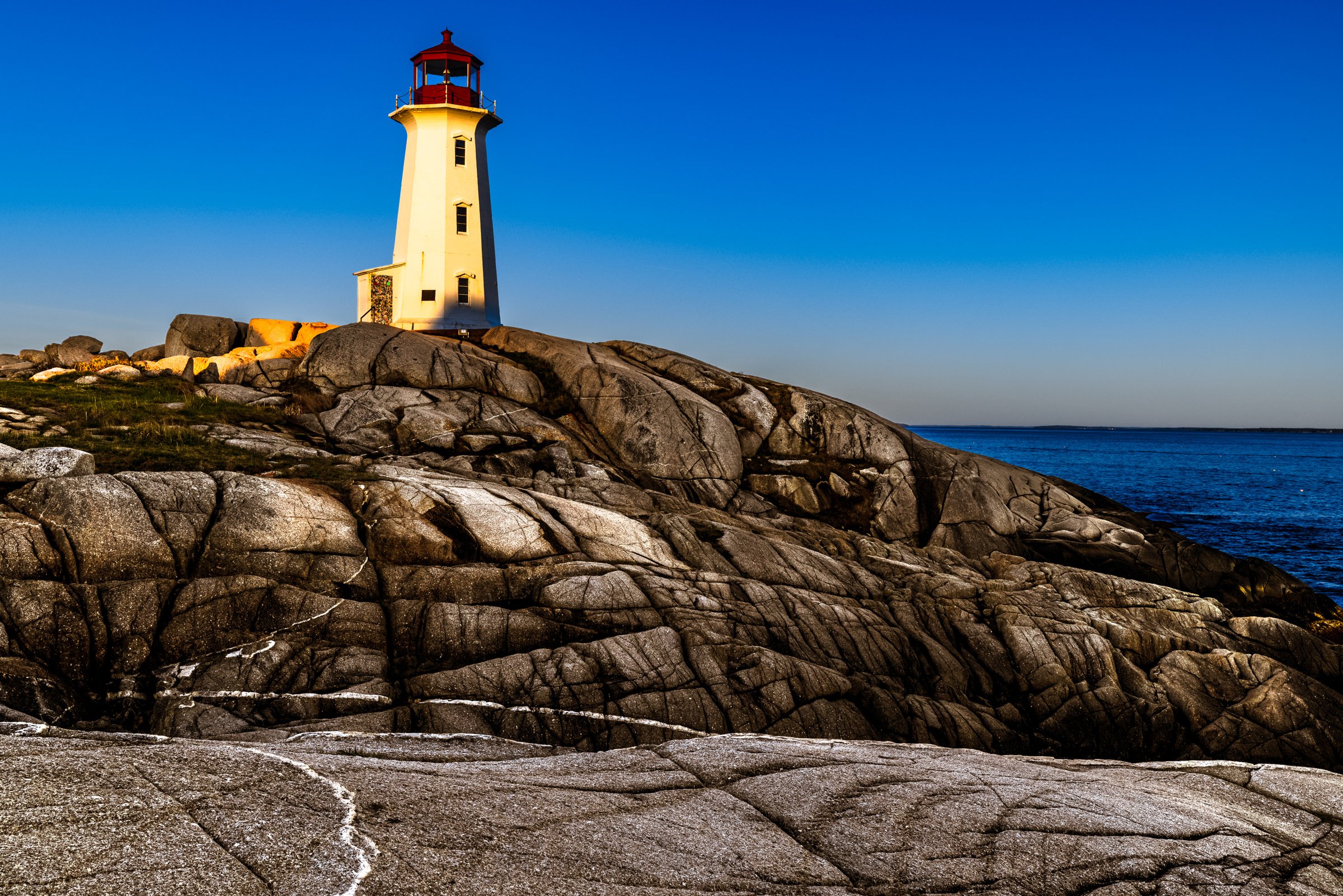 A white lighthouse with a red roof stands atop rugged, gray granite rocks. The structure is bathed in warm sunlight, casting shadows on the textured stones. The sky is a clear, vibrant blue, meeting the deep blue ocean in the distance. The scene conveys a sense of tranquility and isolation, typical of a coastal landscape.