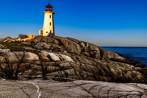A white lighthouse with a red roof stands atop rugged, gray granite rocks. The structure is bathed in warm sunlight, casting shadows on the textured stones. The sky is a clear, vibrant blue, meeting the deep blue ocean in the distance. The scene conveys a sense of tranquility and isolation, typical of a coastal landscape.