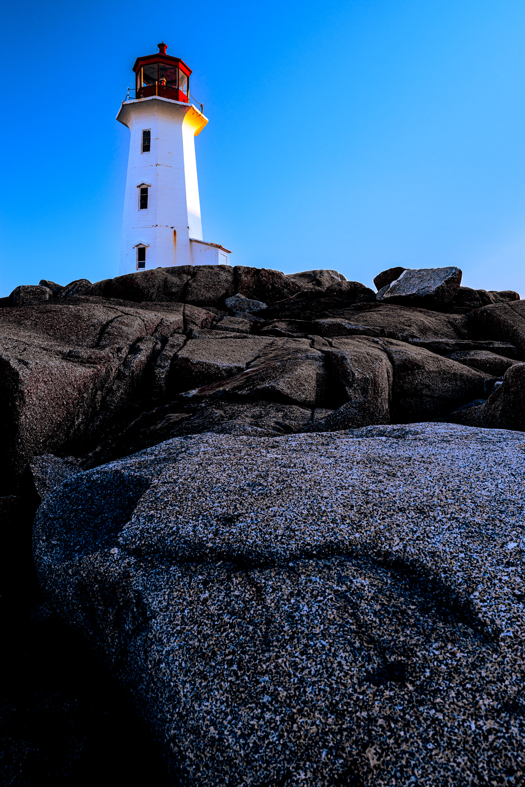 A white lighthouse tower with a red octagonal lantern room stands atop fractured granite boulders at dusk, its gallery light glowing against a deep blue sky.