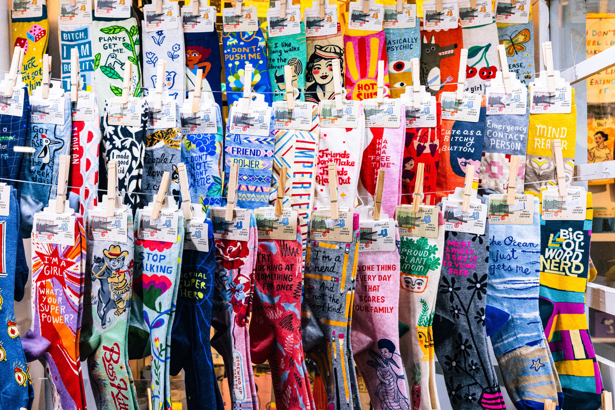 A densely packed retail display of colorful novelty crew socks clipped to horizontal wires with wooden clothespins. The socks feature bold illustrated graphics and humorous text phrases including "Friend Power," "Big Ol' Word Nerd," "Emergency Contact Person," "Stop Talking," "I'm a Girl — What's Your Super Power?," "Basically Psychic," and others. All socks appear to be from the Blue Q brand, visible on hanging header cards. The display fills the entire frame in rows of vivid colors — yellow, red, teal, pink, blue, and gray — against a bright retail background.