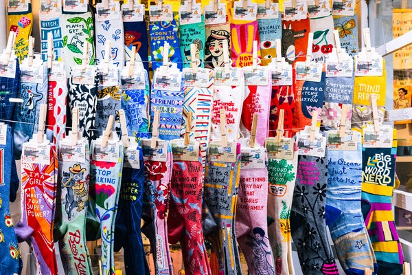 A densely packed retail display of colorful novelty crew socks clipped to horizontal wires with wooden clothespins. The socks feature bold illustrated graphics and humorous text phrases including "Friend Power," "Big Ol' Word Nerd," "Emergency Contact Person," "Stop Talking," "I'm a Girl — What's Your Super Power?," "Basically Psychic," and others. All socks appear to be from the Blue Q brand, visible on hanging header cards. The display fills the entire frame in rows of vivid colors — yellow, red, teal, pink, blue, and gray — against a bright retail background.