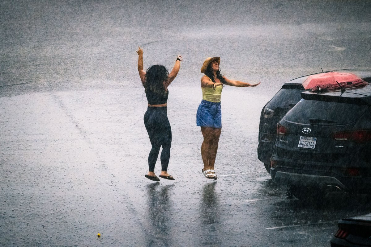 Two women dancing in heavy rain on a wet street along side a row of parked cars. One wears a black outfit and raises her arms joyfully. The other, in a yellow top and blue shorts, wears a straw hat and extends her arms. A black SUV with a red umbrella extending partially over its roof is parked nearby, presumably waiting for the women to finish their impromptu celebration of being caught in a rain storm. The rain creates a blurred, misty background, emphasizing the lively, carefree moment.