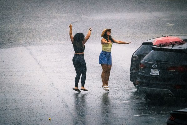 Two women dancing in heavy rain on a wet street along side a row of parked cars. One wears a black outfit and raises her arms joyfully. The other, in a yellow top and blue shorts, wears a straw hat and extends her arms. A black SUV with a red umbrella extending partially over its roof is parked nearby, presumably waiting for the women to finish their impromptu celebration of being caught in a rain storm. The rain creates a blurred, misty background, emphasizing the lively, carefree moment.