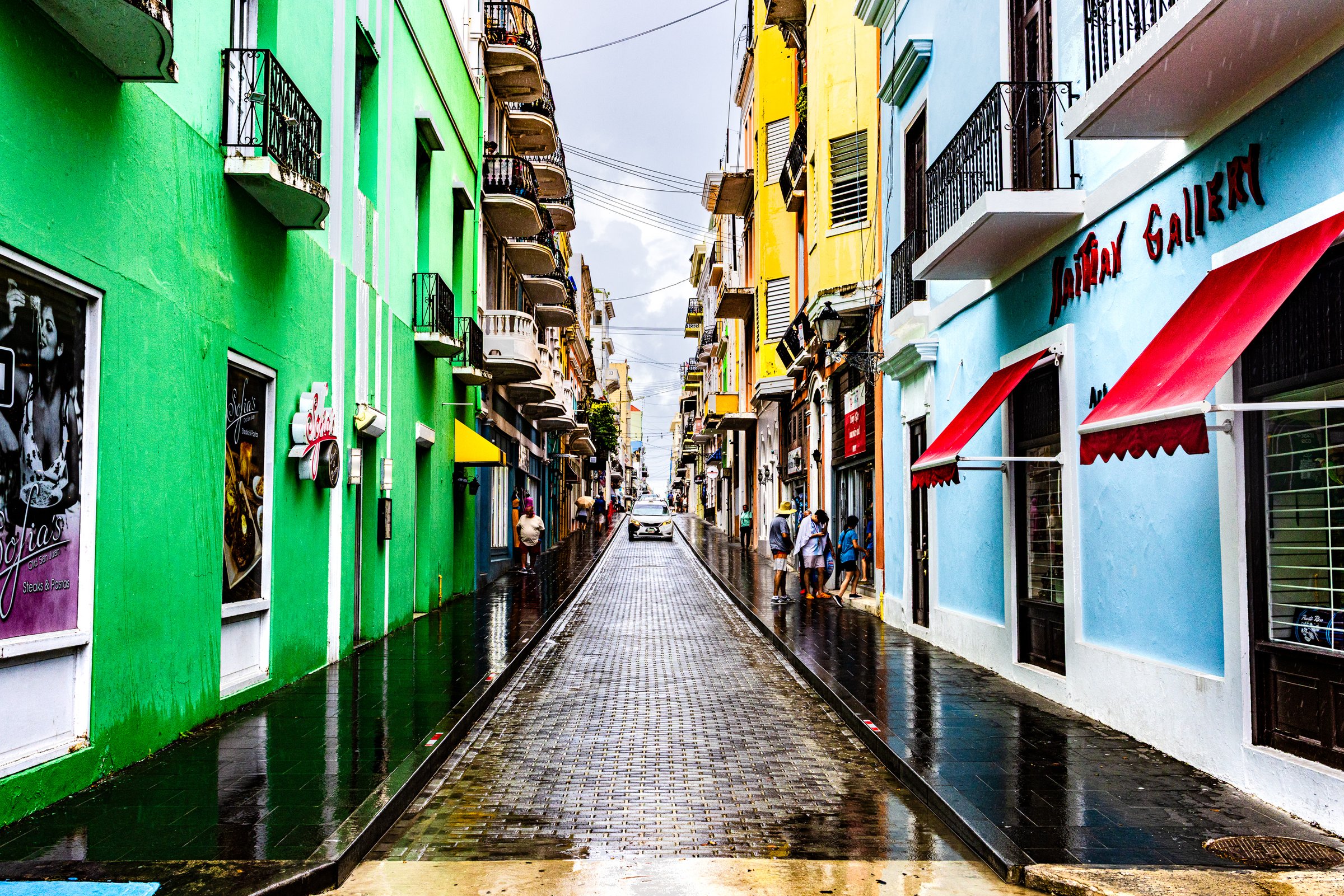 A narrow, cobblestone street flanked by vibrant buildings in bright green, yellow, and blue. Wet pavement reflects the colorful facades and a cloudy sky. Red awnings adorn the right side, housing a gallery. People walk along the sidewalks, and a car is visible in the distance. The scene conveys a lively, urban atmosphere with a touch of rain.