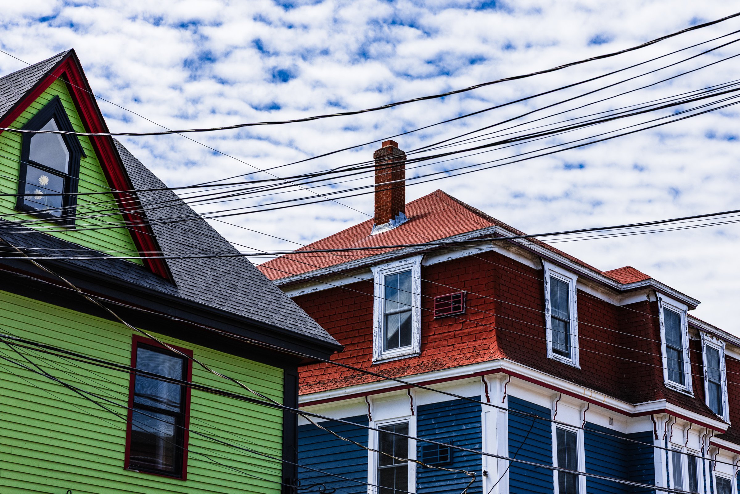 Two colorful houses under a partly cloudy sky. The house on the left is bright green with a black roof and a triangular window. The house on the right is red with a blue lower section and white trim. A brick chimney rises from the red house. Numerous black power lines crisscross the sky above the houses.