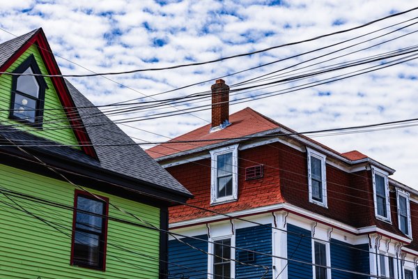 Two colorful houses under a partly cloudy sky. The house on the left is bright green with a black roof and a triangular window. The house on the right is red with a blue lower section and white trim. A brick chimney rises from the red house. Numerous black power lines crisscross the sky above the houses.